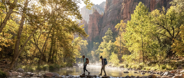Two hikers with backpacks crossing Oak Creek on stepping stones beneath red rock cliffs and golden fall cottonwoods near Sedona