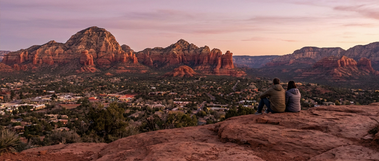 Couple sitting on a red rock ledge at the Airport Mesa overlook watching the sunset over Sedona, Arizona and its red rock buttes